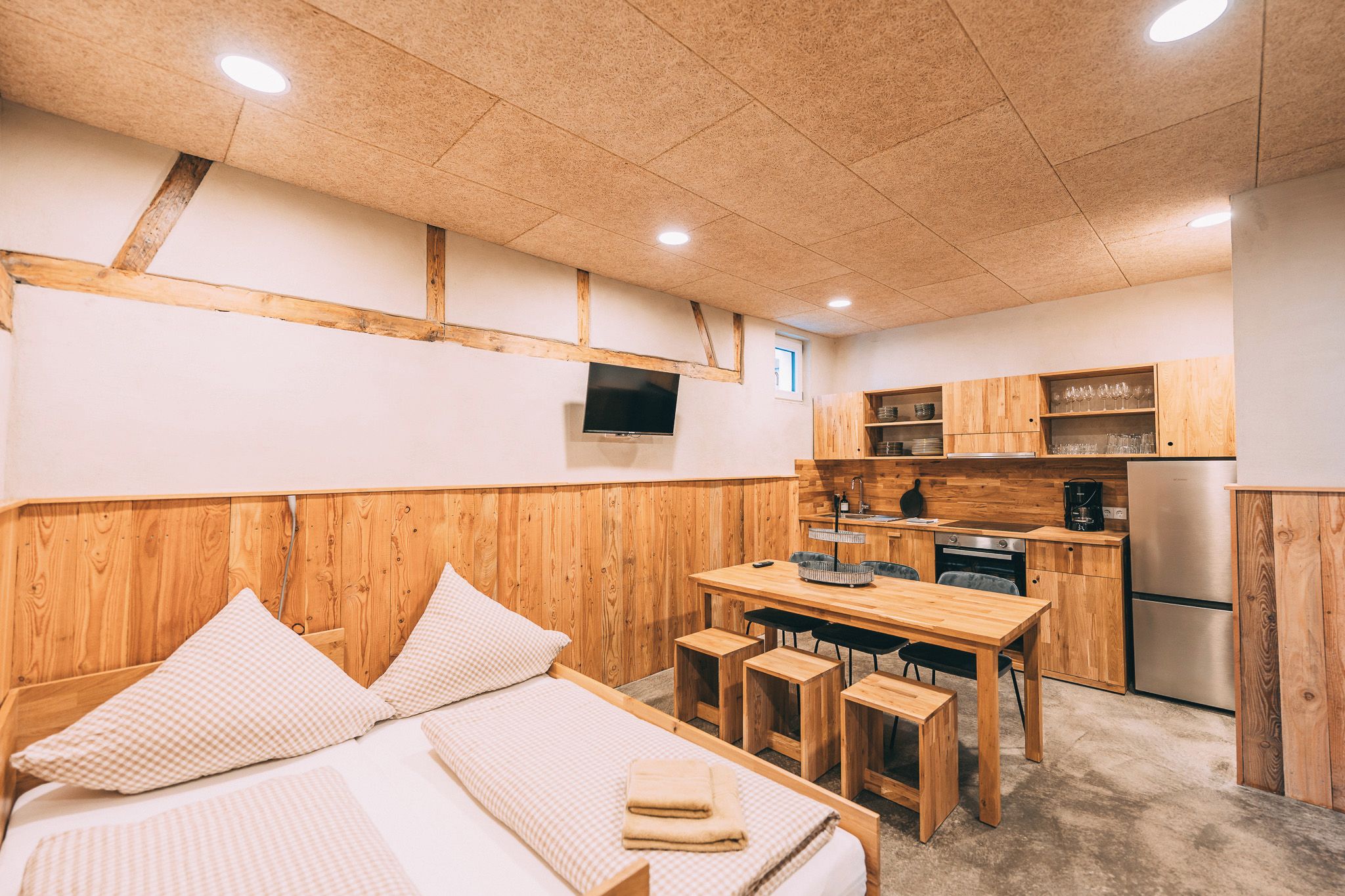Een knus verblijf met een bed, een houten eettafel, een kitchenette met een koelkast en een televisie aan de muur.