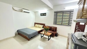 A hotel room with a bed, a small table with a vase of flowers, a sofa, a window with blinds, and an air conditioning unit. - Era Apartment Kham Thien (Hanoi)