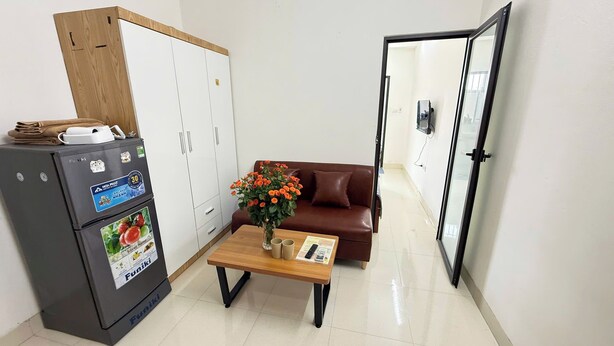 A compact living space with a kitchenette, a brown leather sofa, a small coffee table with a vase of flowers, and a door leading to another room. - Era Apartment Kham Thien (Hanoi)