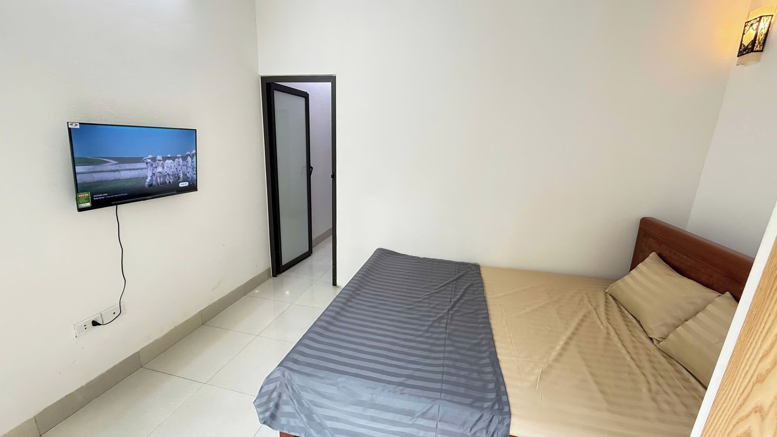 A small, clean room with a bed, a TV mounted on the wall, and a door leading to another room.