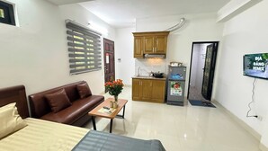 A compact living space with a sofa, a small table with a vase of flowers, a kitchenette with a refrigerator and stove, and a TV mounted on the wall. - Era Apartment Kham Thien (Hanoi)