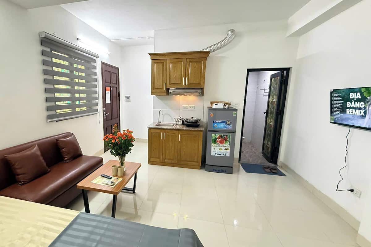 A compact living space with a sofa, a small table with a vase of flowers, a kitchenette with a refrigerator and stove, and a TV mounted on the wall.