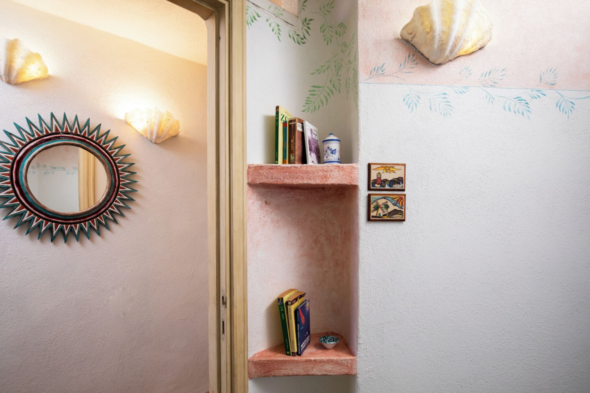 A bathroom with a sunburst mirror, shelves with books and a small bowl, and decorative shells on the wall.