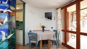 Un comedor con una mesa, sillas, un florero con flores, un televisor en la pared y vistas a través de una puerta de vidrio.