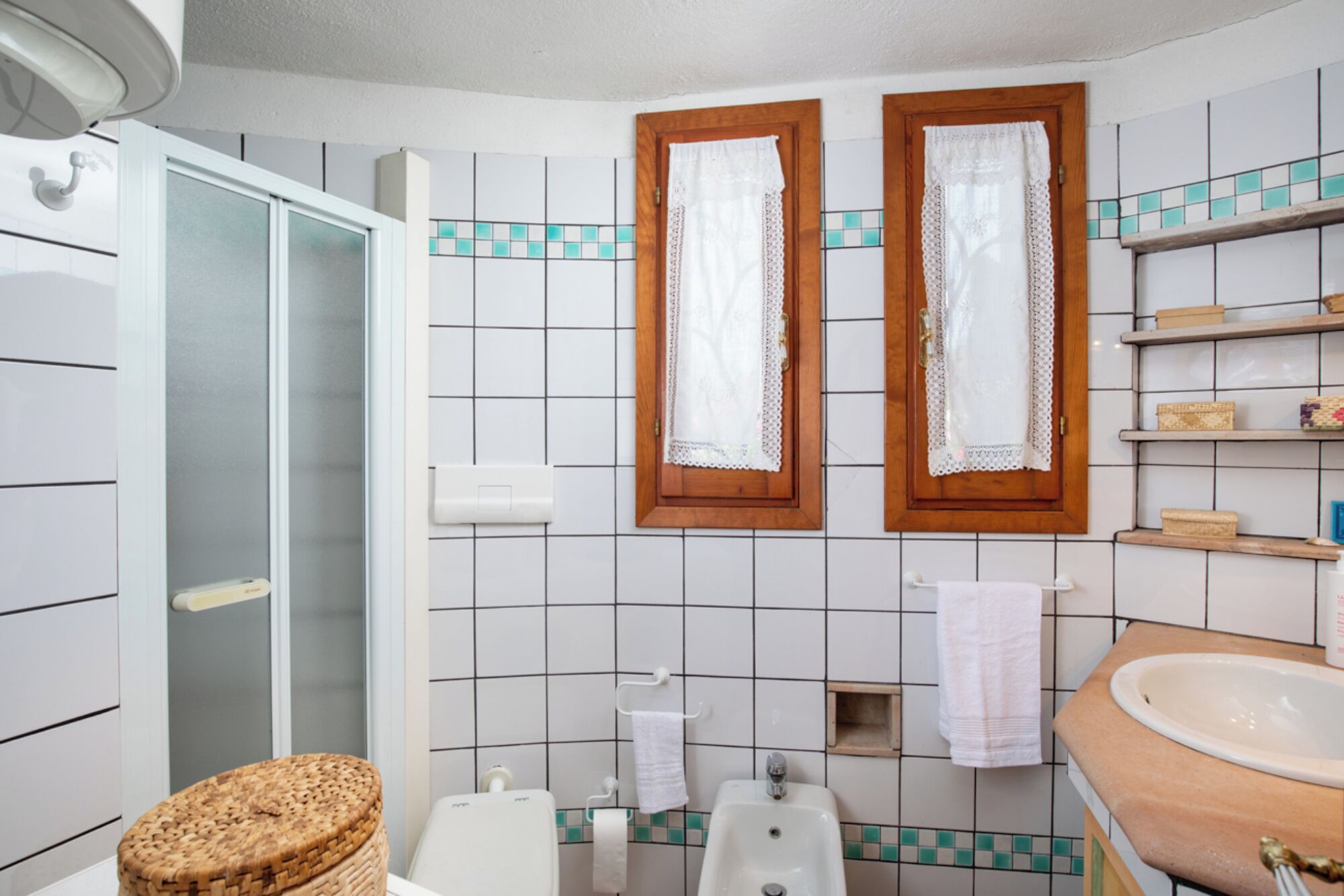 A bathroom with a shower, a sink, and a toilet. There are two windows with wooden frames and lace curtains.