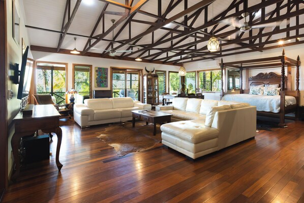 A spacious room with a high ceiling, wooden beams, a large bed with a canopy, a sofa, a coffee table, and a television.