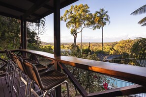 A covered outdoor seating area with wicker chairs and a wooden railing overlooking a forested landscape.