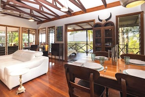 A spacious living area with a white sofa, wooden dining table set for four, and a view of the outdoors through large windows. - Mount Panorama Port Douglas Ocean and valley views Tropical Oasis Pool Garden (Port douglas)
