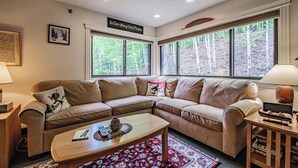 A cozy living room with a sectional sofa, a wooden coffee table, and a large window with a view of trees.