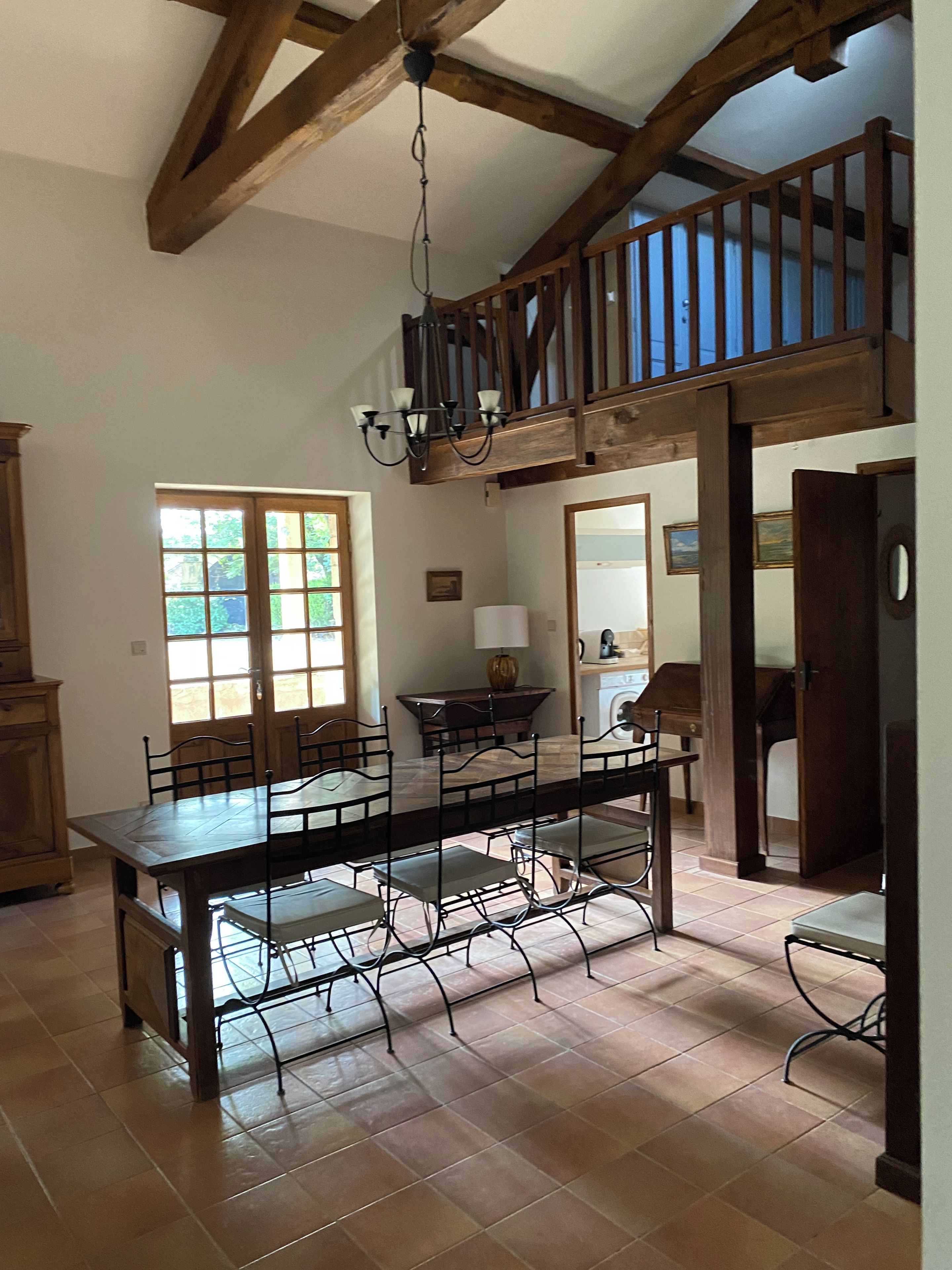 A spacious dining area with a high ceiling and exposed wooden beams, a chandelier, and a view of the outdoors through a window.