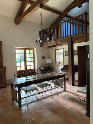 A spacious dining area with a high ceiling and exposed wooden beams, a chandelier, and a view of the outdoors through a window.