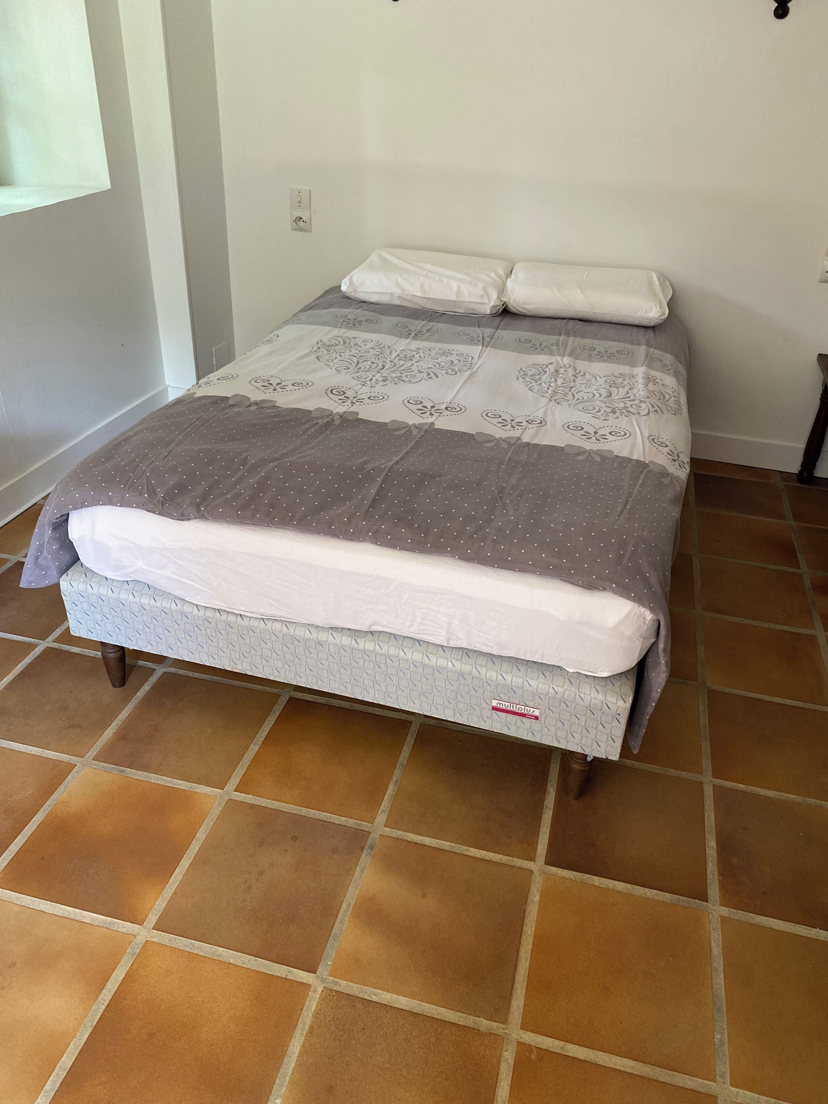 Ein Bett mit einem gemusterten Überwurf, einem weißen Kopfkissen und einer Matratze mit der Aufschrift 'multiplex'. Der Raum hat einen Fliesenboden und weiße Wände.