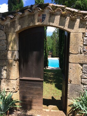 A stone archway leading to a wooden door, with a view of a swimming pool and greenery in the background.