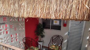 A room with a thatched roof, two wicker chairs with floral cushions, a small table, and a red wall. - CHALES DO TOQUE (São Miguel dos Milagres)