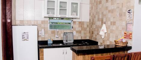 Een kleine keuken met witte kastjes, een zwart aanrecht, een ingebouwde magnetron en een houten stoel.