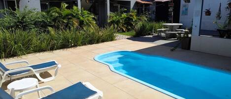 Una piscina con sillas de descanso y una pequeña mesa, rodeada de vegetación y un edificio con balcones.
