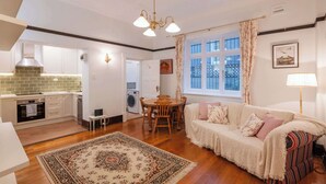 A modern kitchen with white cabinets, a dining area with a wooden table and chairs, a cozy living room with a sofa and a patterned rug, and a large window with curtains.