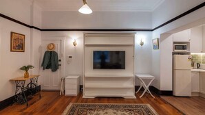 A room with a flat-screen TV mounted on a white wall, a small white table, a white cabinet, and a wooden floor.