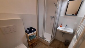 A bathroom with a toilet, a shower enclosure, a sink, and a small storage shelf. - Kirchenwirt Halbwax (Kaumberg)