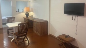 Une petite pièce bien éclairée, avec un bureau, une chaise, une lampe, un téléviseur fixé au mur et une télécommande posée sur une petite table.