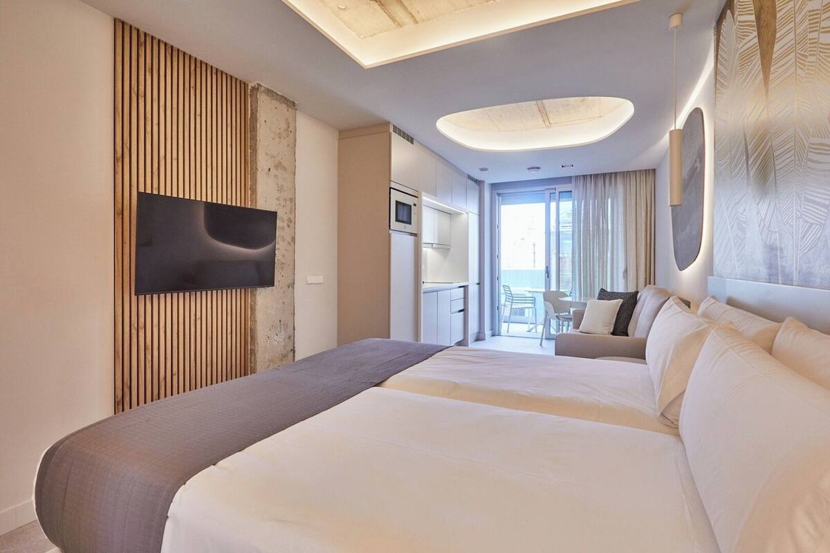 A hotel room with a large bed, a TV mounted on the wall, a kitchenette, and a balcony with a view.