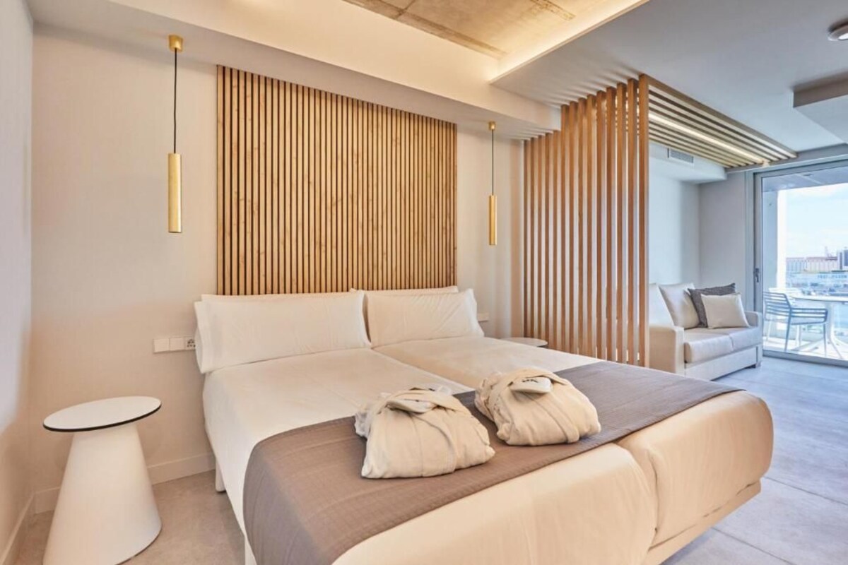 A modern hotel room with a large bed, a small side table, a sofa, and a view of the city through large windows.