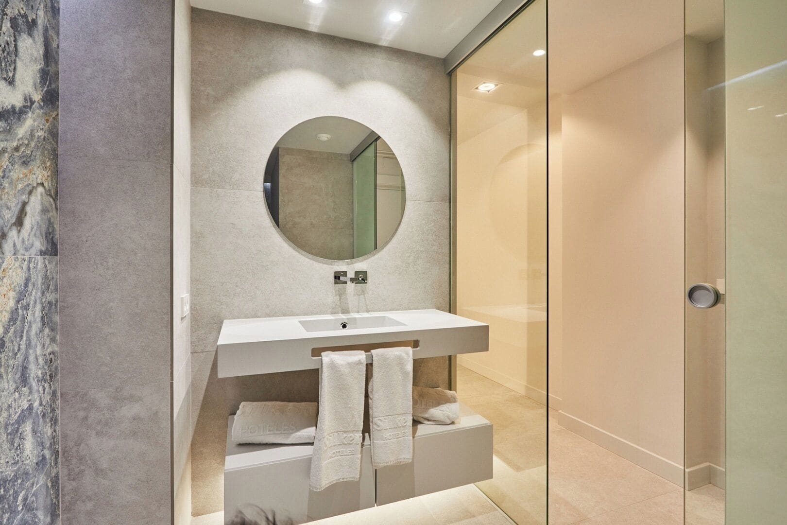 Un baño moderno con pared de mármol, un espejo grande, un lavabo blanco y una cabina de ducha de vidrio.