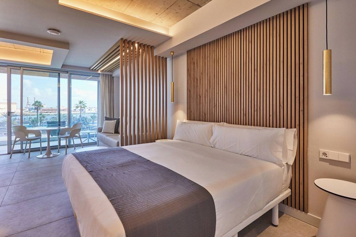 A modern hotel room with a large bed, a balcony with a table and chairs, and a view of the cityscape.