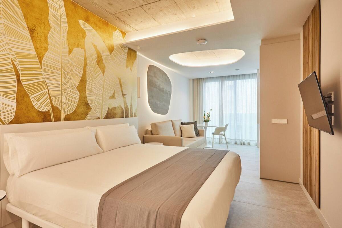 A modern hotel room with a large bed, a seating area with a sofa and chairs, a flat-screen TV mounted on the wall, and a large window with curtains.