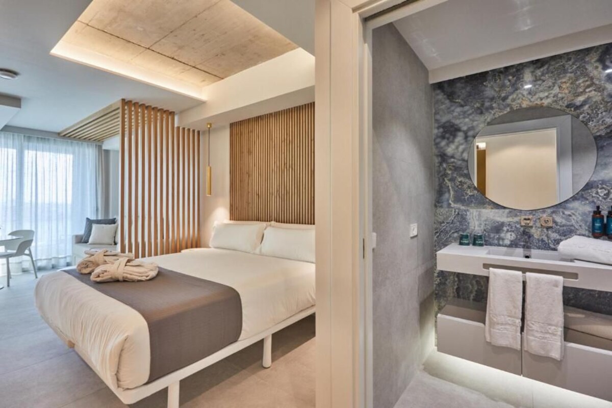 Een moderne hotelkamer met een bed, een wand van houten panelen, een marmeren wastafel in de badkamer en een ronde spiegel.