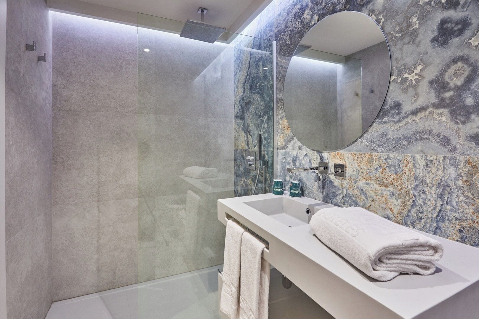 Une salle de bain moderne avec une douche à parois de verre, un meuble-lavabo blanc doté d'un miroir rond et des murs à motifs de marbre.