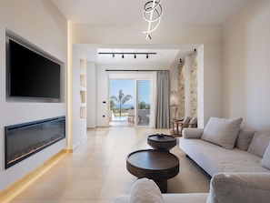 A modern living room with a flat-screen TV, a fireplace, a sofa, and a view of the ocean through a sliding glass door.