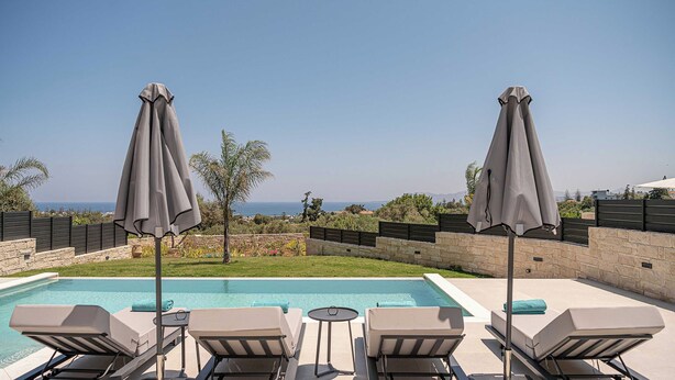 A poolside area with lounge chairs and umbrellas, a small table, and a view of the sea. - Kymélia Ground Suite With Private Pool & SeaView (Galatas)