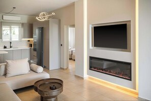 A modern living room with a fireplace, a sofa, a coffee table, and a kitchen area with a refrigerator and a flat-screen TV. - Kymélia Ground Suite With Private Pool & SeaView (Galatas)