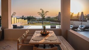 A wooden table with a fruit bowl, surrounded by wicker chairs, overlooking a pool area with lounge chairs and umbrellas. - Kymélia Ground Suite With Private Pool & SeaView (Galatas)
