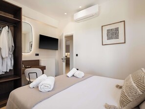 A hotel room with a bed, a bedside table, a chair, a wardrobe, a mirror, a TV, and an air conditioning unit. - Kymélia Ground Suite With Private Pool & SeaView (Galatas)
