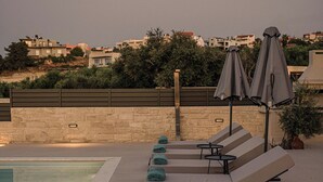 Uma área de piscina no terraço com espreguiçadeiras e guarda-sóis, piscina cristalina e vista para edifícios ao fundo.