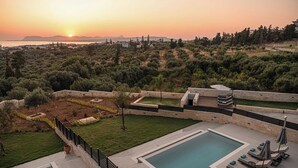 Un espace piscine comprenant deux piscines distinctes, des chaises longues et des parasols, le tout entouré d’un jardin aménagé et offrant une vue sur le coucher de soleil sur la mer.