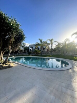 Una piscina de borde curvo, rodeada por un patio empedrado. Se ven palmeras y un edificio de piedra con arcos al fondo.