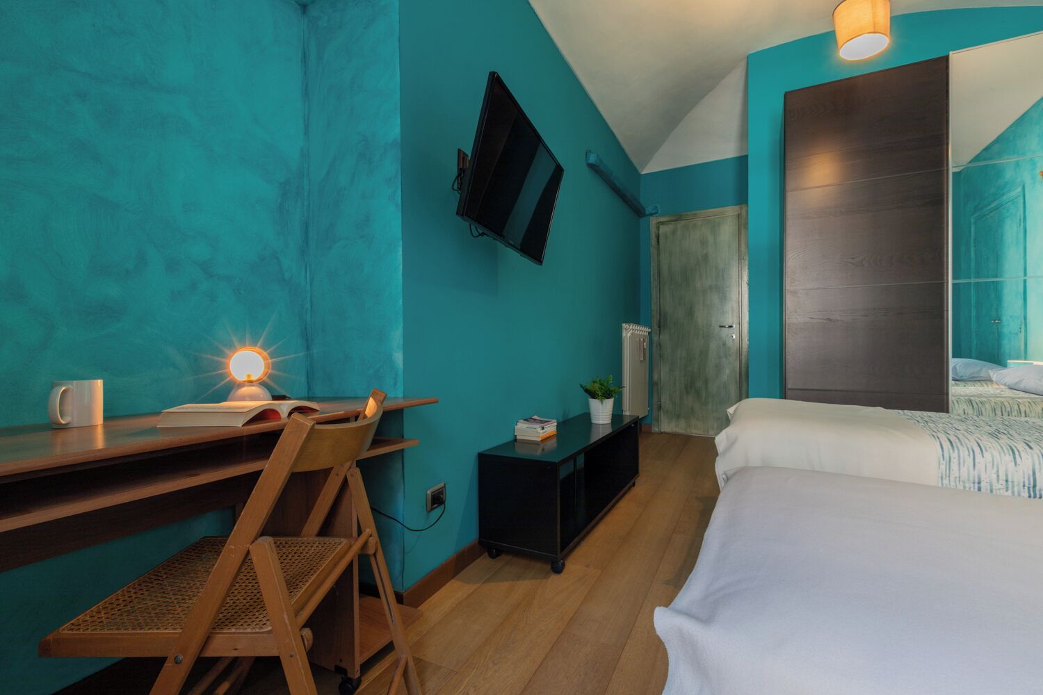 Una habitación con paredes color turquesa, un escritorio de madera con silla, una cama con ropa de cama blanca, un televisor montado en la pared y una mesita con una planta.