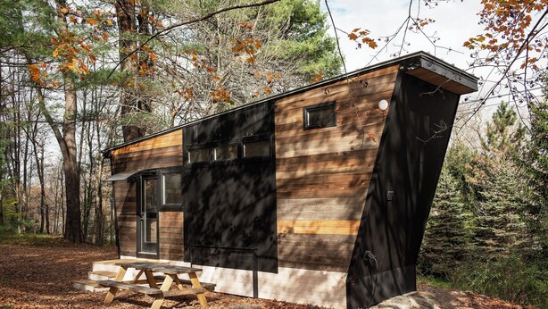 A small cabin with a sloped roof, a front door, and a picnic table in a wooded area.