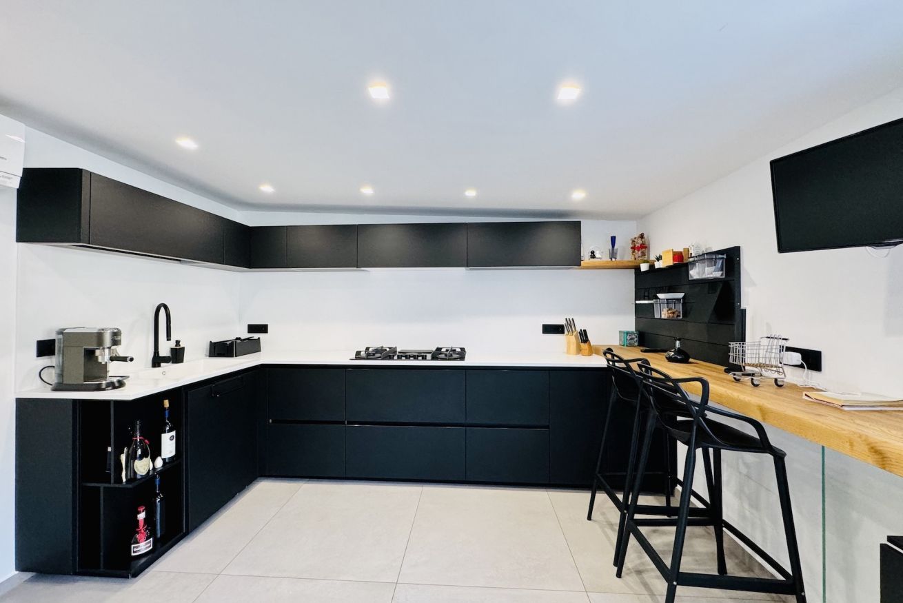 A modern kitchen with black cabinets, a white countertop, a coffee machine, and a TV mounted on the wall.