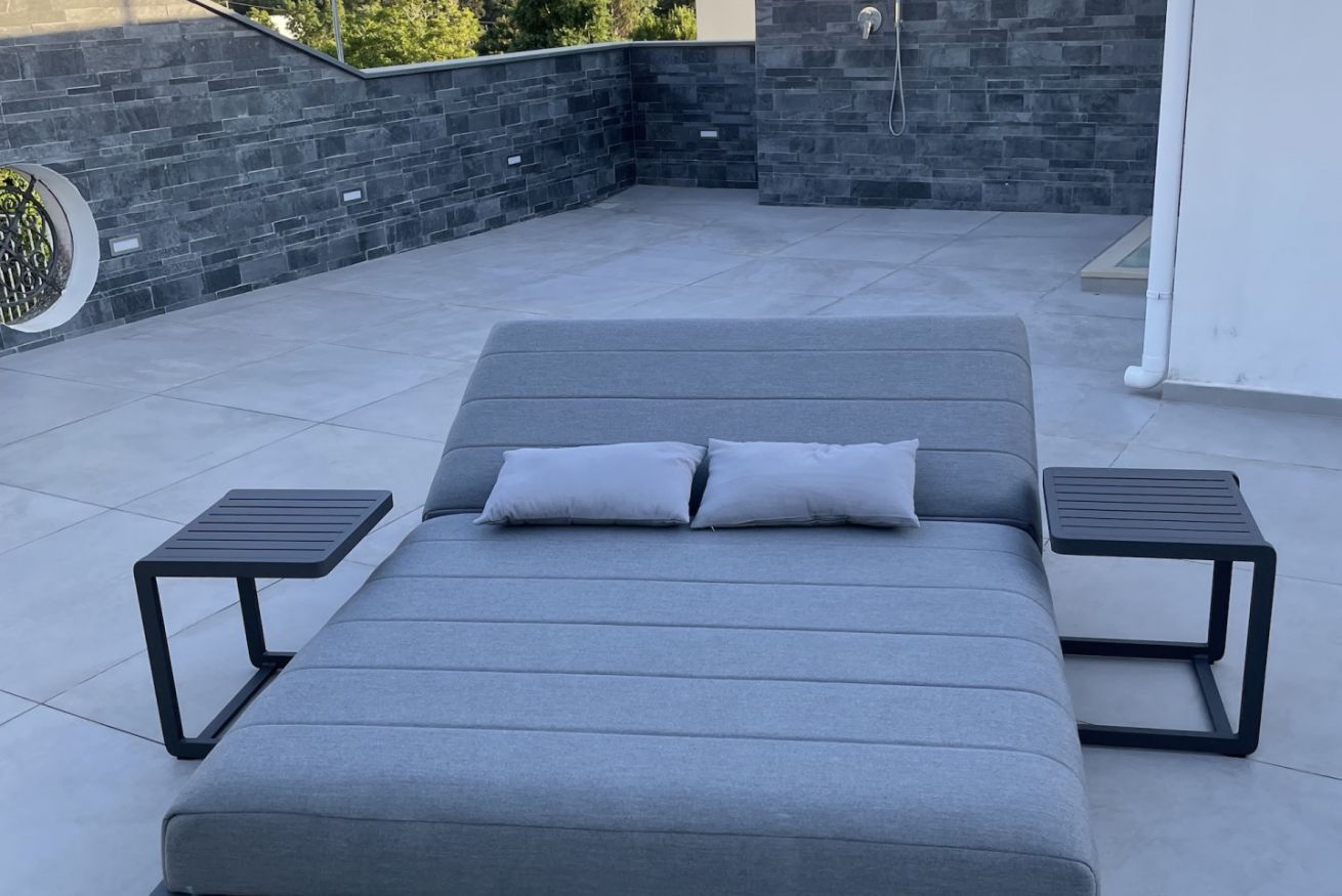 A modern outdoor seating area with a grey sofa, two side tables, and a stone wall backdrop.