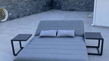 A modern outdoor seating area with a grey sofa, two side tables, and a stone wall backdrop.
