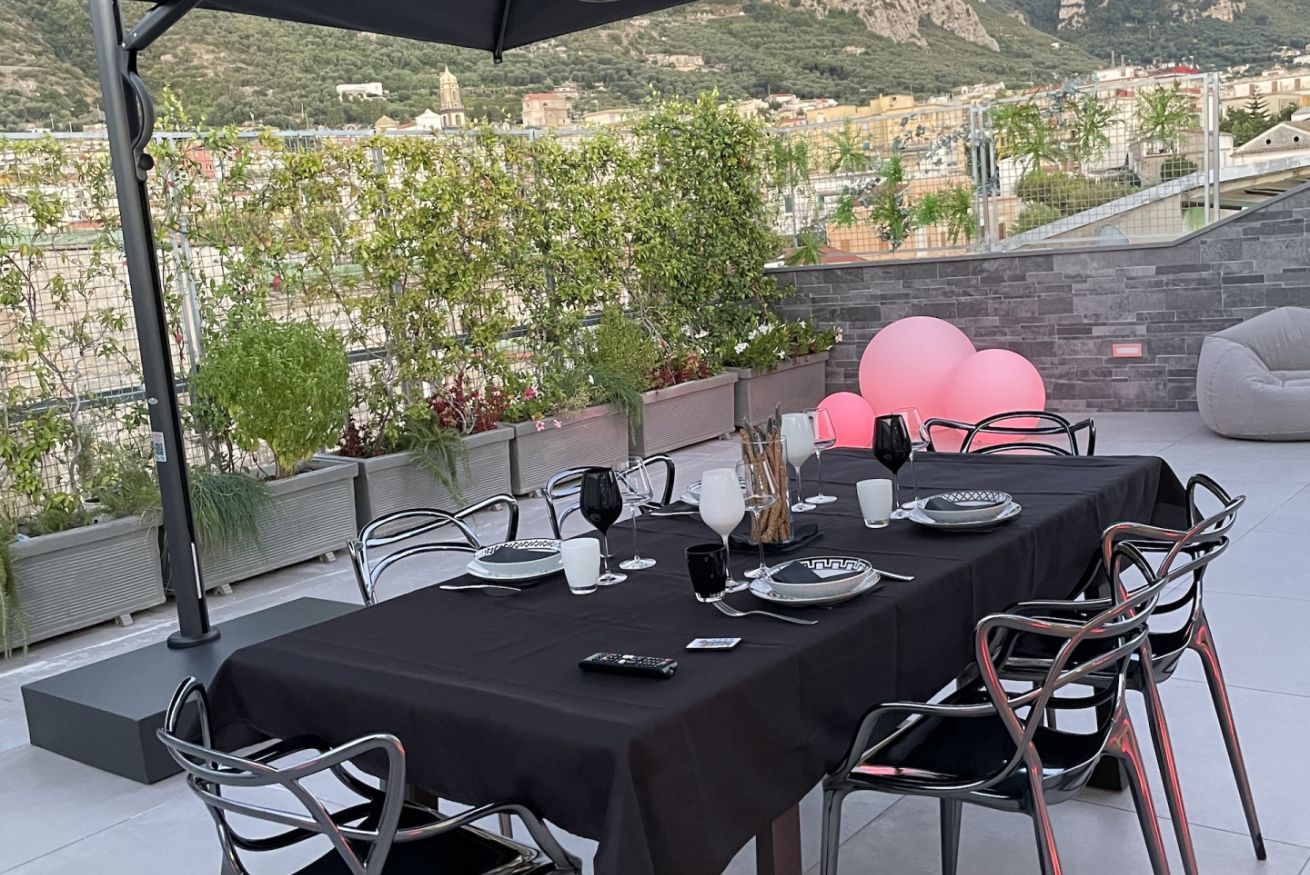 Un área de comedor al aire libre con una mesa dispuesta para cuatro personas, rodeada de sillas de metal, y una vista panorámica de la ciudad de fondo.
