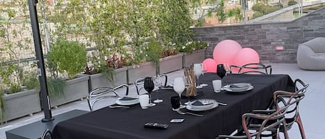 Un área de comedor al aire libre con una mesa dispuesta para cuatro personas, rodeada de sillas de metal, y una vista panorámica de la ciudad de fondo.