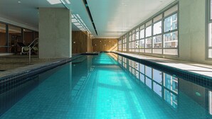 Una larga piscina interior con agua cristalina azul, grandes ventanales en un lado y una escalera en el otro.