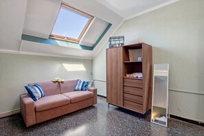 Een kamer met een schuine kap, een raam, een bruine bank met kussens, een houten kast en een hoge spiegel.