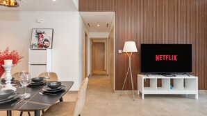A modern living room with a flat-screen TV displaying 'NETFLIX', a dining area with a table set for four, and a hallway leading to another room.