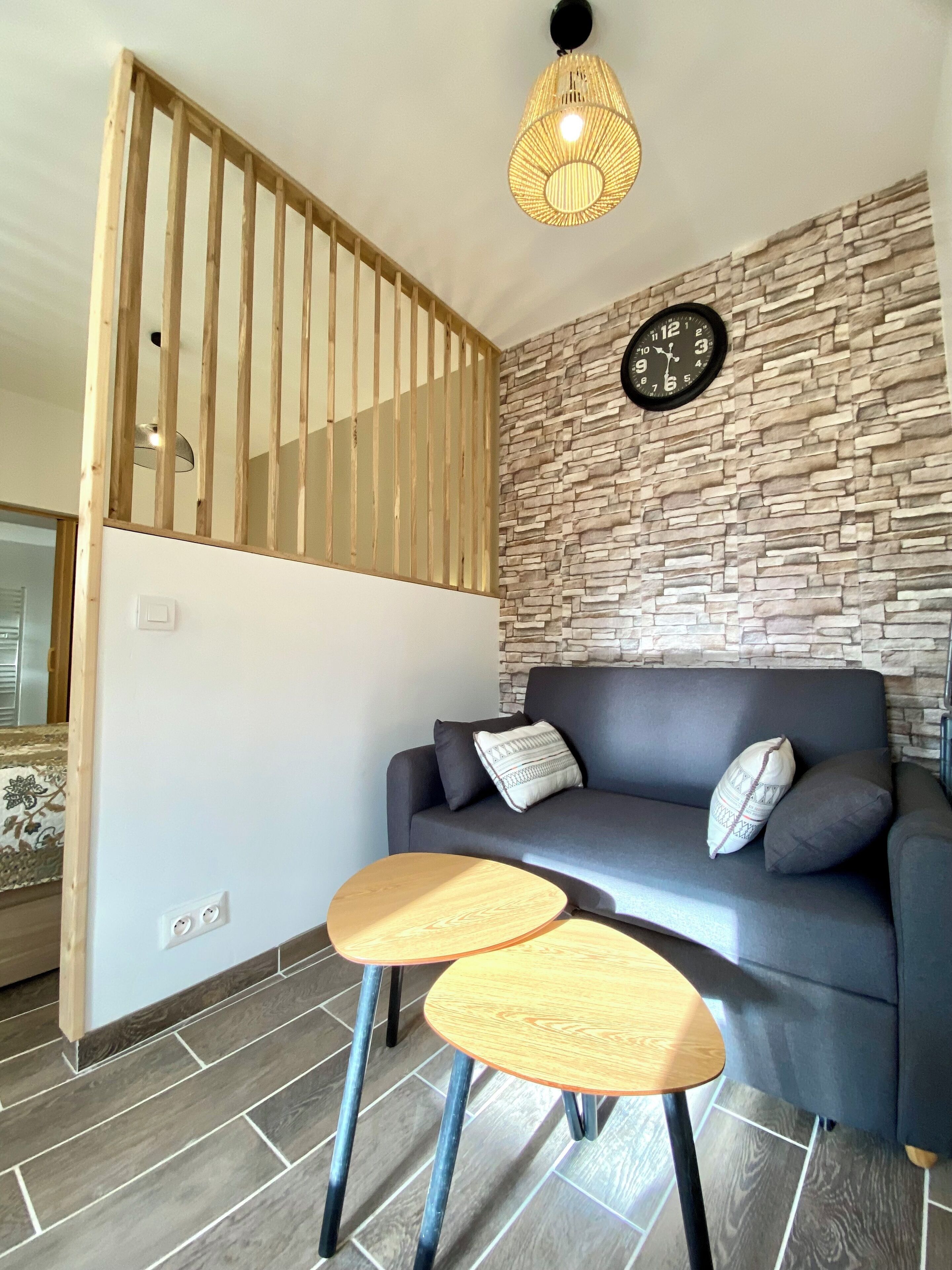 A modern living room with a grey sofa, two wooden side tables, a stone wall, a wooden partition, and a hanging light fixture.